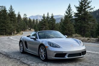 Porsche Boxster argentée garée sur une route de montagne sinueuse, vue en angle 3/4 avant avec forêt de pins en arrière-plan