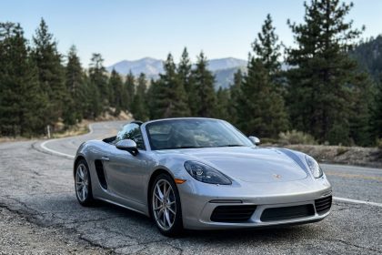 Porsche Boxster argentée garée sur une route de montagne sinueuse, vue en angle 3/4 avant avec forêt de pins en arrière-plan